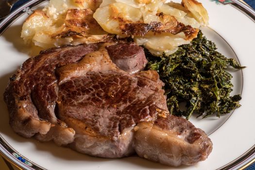 Large grilled ribeye steak on white plate with roasted kale and sliced potatoes