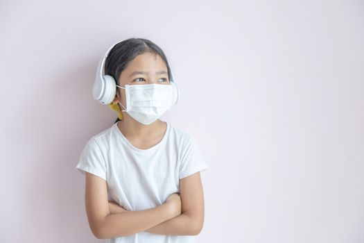 A little Asian girl wearing a medical protective mask and earphones. The child girl standing cross one's arm on the color background. The epidemic of the flu, Coronavirus or COVID-19 and illness concept.