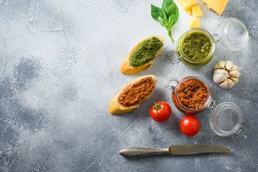 Glass jars with red and green pesto and cooking italian recipe ingredients Parmesan cheese, basil leaves, pine nuts, olive oil, garlic, salt, tomatoes breakfast panini with sauce top view on grey concrete surface space for text