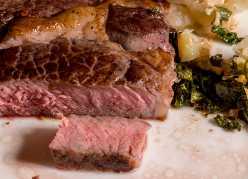 Large grilled ribeye steak with slice on white plate with roasted kale and sliced potatoes