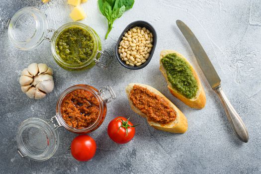 Glass jars with red and green pesto and cooking italian recipe ingredients Parmesan cheese, basil leaves, pine nuts, olive oil, garlic, salt, tomatoes breakfast panini with sauce top view on grey concrete surface space for text.