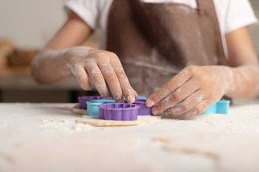 A little girl is wearing a brown apron using a purple mold to cut the dough for making cookies in the kitchen.