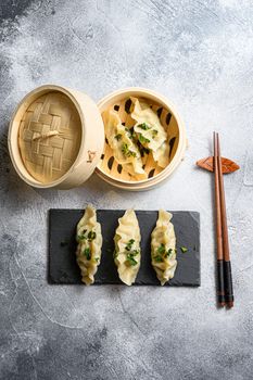 Dim sum dumplings in steamer and on a stone plate with chopsticks and fresh herbs top view on grey background overhead.