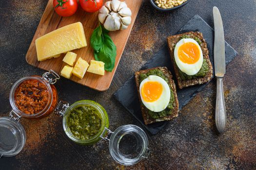 Glass jars with red and green pesto and panini bread with pesto and eggs breakfast italy style cooking ingredients Parmesan cheese, basil leaves, pine nuts, white stone background, close up top view
