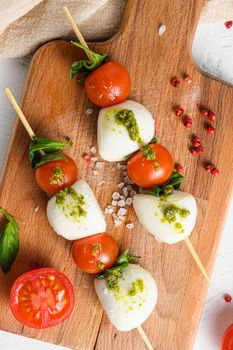 Caprese salad on sticks Cherry tomatoes, mozzarella cheese, basil, pesto sauce on wood board over white rustic background top view close up.