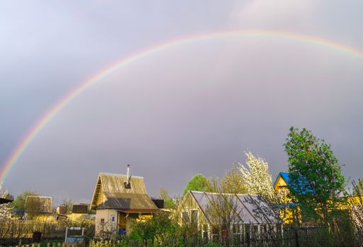 Rainbow over the roofs of rural houses in the stormy evening sky after a storm, summer village landscape.