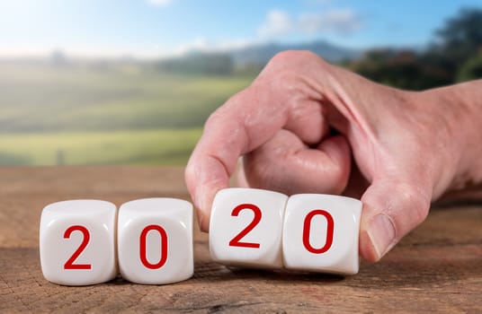 2020 spelled in cubes with man's hand turns over the dice on wooden table with sunlight on rural landscape