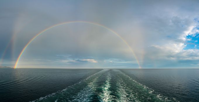 Colorful and dramatic double rainbow forms a full arch over the wake of a departing cruise ship at sea