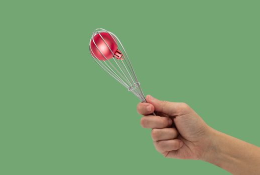 Female hand with a kitchen whisk and ball christmas isolated on green background.