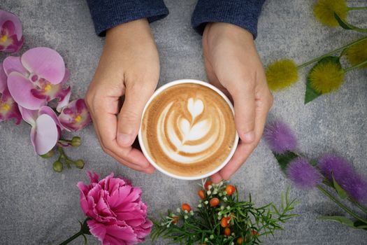 The hands in the sweater are holding a cup of coffee and there are many colored flowers surrounded.