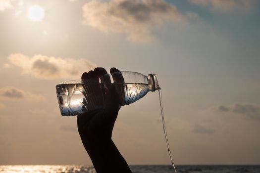 The hand is pouring refreshing water from the plastic bottle on the beach at evening time.