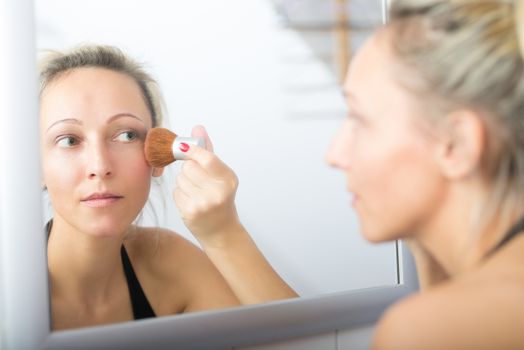A blonde woman powdering her skin, photographed in the mirror