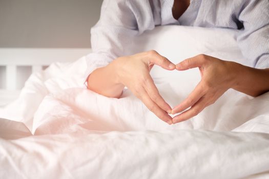 Woman is showing a gesture by hand in heart shape on the white mattress in the morning, with the orange light from the sun shining.