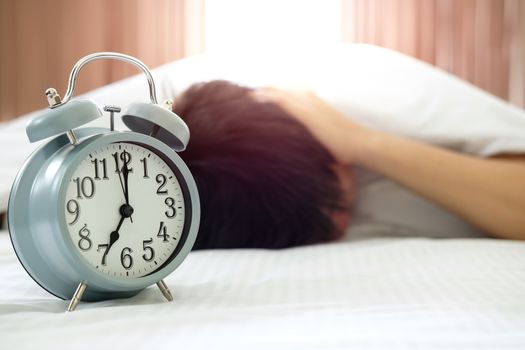 Woman covering ears with pillow in bed with alarm clock
