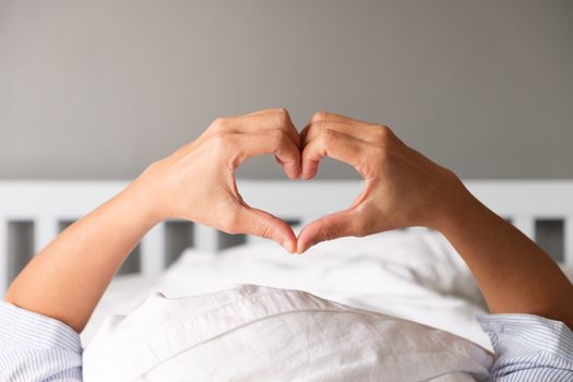 Woman is showing a gesture by hand in heart shape on the white mattress in the morning.