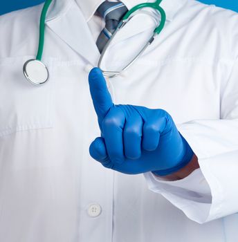 male doctor in a white medical coat and blue latex gloves stands on a blue background and with his right hand stretches his index finger forward, an imitation of pressing