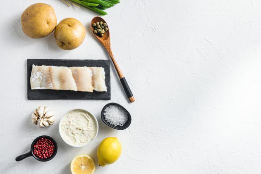 Traditional fish and chips ingredients recipe raw cod fillets on stone slate batter, potatoe, tartar sauce, lemon, capers , green herbs garlic, salt, peppercorns on white stone background flat lay top view space for text.