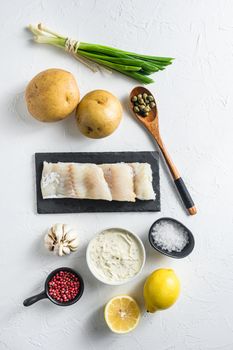 Traditional fish and chips ingredients recipe raw cod fillets on stone slate batter, potatoe, tartar sauce, minty mushy peas, lemon , shallot, mint, garlic, salt, peppercorns on white stone background flat lay top view .
