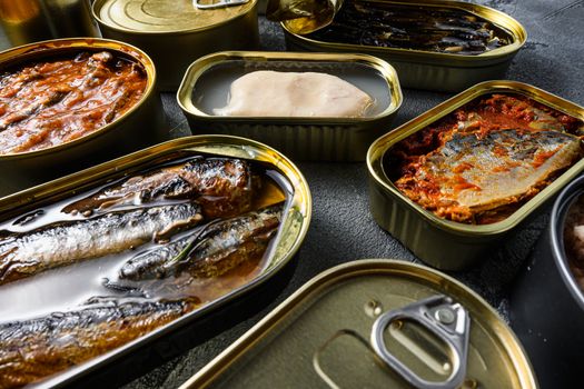 Conserves of canned fish with different types of fish and seafood, opened and closed cans with Saury, mackerel, sprats, sardines, pilchard, squid, tuna, over grey stone surface, side view close up new widw angle.