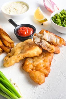 Fishand chips battered crispy cod fillet detail with dip and lemon in a paper cone on white background with all components classic recipe takeaway food white stone textured background side view vertical photo.