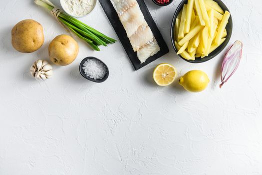 Traditional fish and chips ingredients recipe raw cod fillets on stone slate batter, potatoe, tartar sauce, lemon, capers , green herbs garlic, salt, peppercorns on white stone background top view concept in raw space for text on bottom.