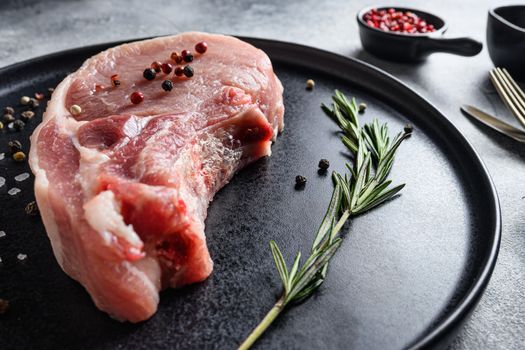 Raw Pork Loin chops in a black round plate on a grey textured stone background with rosemary garlic peppercorns ingredients for grill close up selective focus new wide angel.