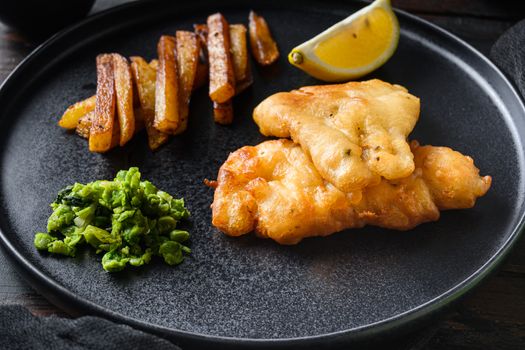 Fish and chips. Traditional british hot dish consisting of fried fish, potato chips, mushy peas and tartare sauce. served on black plate over old dark wood table in pub side view.