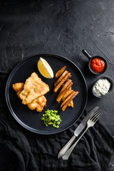 Fish and chips. Traditional british hot dish fried fish, potato chips, mushy minty peas and tartare sauce and ketchup view from above, black stone background space for text.