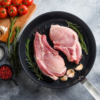 Pork steak on a bone in a black frying grill pan on a grey textured stone background with black cloth rosemary garlic peppercorns and tomatoes on chopping board ingredients top view square .