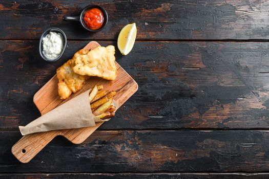 Fish and chips in a paper cone on wood chopping board dip and lemon - fried cod, french fries, lemon slices, tartar sauce, ketchup tomatoe and with mushy peas served in the Pub or Restaurant over old wooden planks dark table top view space for text or recipe