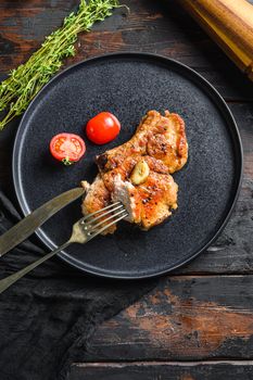 Dish of grilled pork chop with tomatoes top view with knife and slice on fork over old rustic dark wood table table flatlay top view vertical.