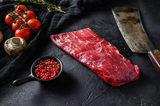 Raw, flap or flank, also known Bavette steak near butcher knife with pink pepper and rosemary. Black stone background. Side view vertical.