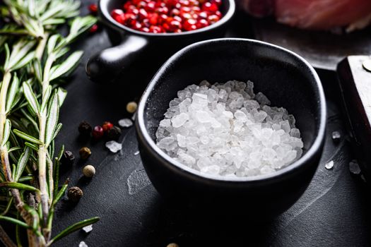 Sea Salt in black bowl and Rosemary herbs close up on black stone table with spices and raw meat near side view selective focus.