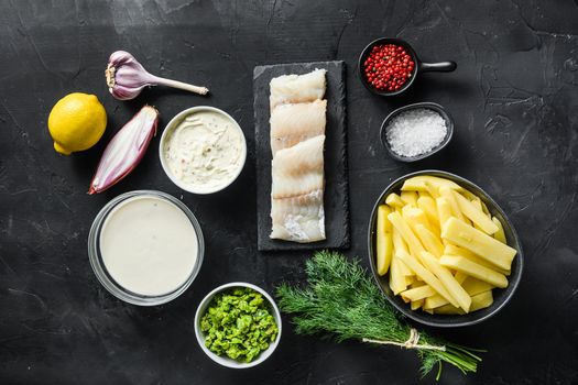 Organic fish and chips ingredients raw cod fillets on stone slate batter, potatoe, tartar sauce, minty mushy peas, lemon , shallot, mint, garlic, salt, peppercorns on black stone background overhead photo.