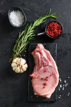 Porco Iberico French Rackswith salt and garlic, pepper on black slate stone board with rosemary, and fresh ingredients for grill over black textured background top view, flatlay on a black stone background vertical.