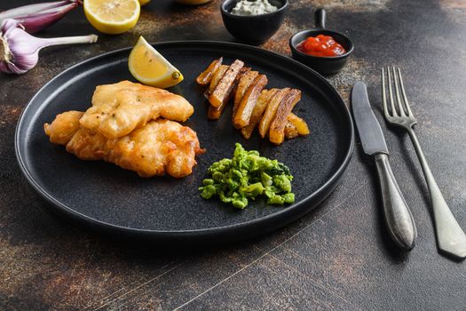 fish and chips with beer-battered cod and fries and a side of tartar sauce and mushy peas on black plate over old rustic metal style background side view new wide angle.