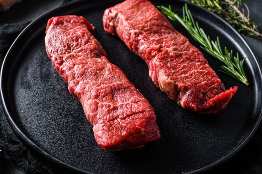 Raw denver cut black angus organic steak on a black plate and stone slate with seasonings, herbs grey concrete background. Side view close up selective focus new wide angle