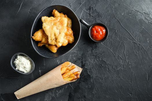 Fish and chips in a paper cone dip and lemon - fried cod, french fries, lemon slices, tartar sauce, ketchup tomatoe and mushy peas over black background top view space for text
