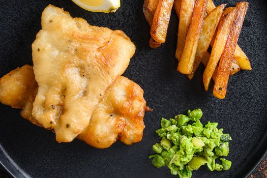 English Traditional Fish and chips with mashed minty peas, tartar sauce vertical side view on black plate close up details side view. Over old rustic table.