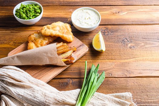 Fish & chips with dip and lemon, mashed minty peas, tartar sauce in paper cone on wood chopping board dip and lemon - fried cod, french fries, lemon slices, tartar sauce, ketchup tomatoe served in the Pub or over light old wooden planks table side view close up space for text.