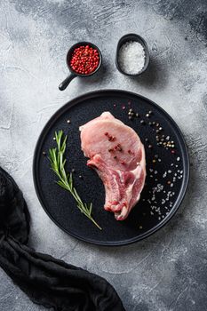 Raw pork chop cutlet in a black round plate on a grey textured stone background with black cloth rosemary garlic peppercorns ingredients top view vertical.