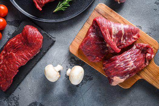 Organic Machete and flank, bavette steak on chopping board, marbled beef over other alternative cuts with herbs tomatoes peppercorns over grey stone surface background top view.