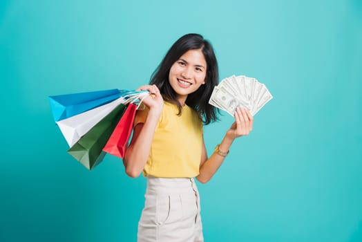 Portrait happy Asian beautiful young woman smile white teeth standing wear yellow t-shirt, She holding shopping bags and dollars money fan, studio shot on blue background with copy space for text