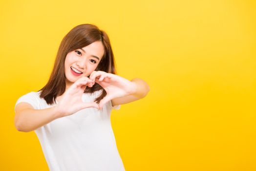 Asian happy portrait beautiful cute young woman teen smile standing make finger heart figure symbol shape sign with two hands looking to camera isolated, studio shot yellow background with copy space