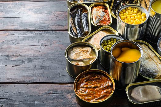 Canned preserves food in tin open cans. with fresh organic ingridients Saury, mackerel, sprats, sardines, pilchard, squid, tuna pinapple, corn, peas, mango , beans, over old wood dark textured background side view space for text.