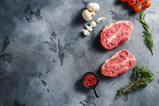 Raw top blade flat Ironcut, on black slate , bio marbled beef with herbs tomatoes peppercorns over grey stone surface background side view space for text.