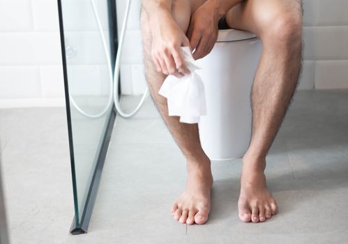 Closeup man sitting on toilet with tissue paper in hand, selective focus