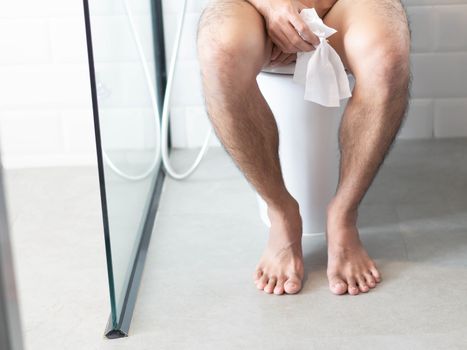 Closeup man sitting on toilet with tissue paper in hand, selective focus