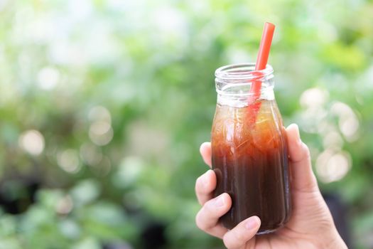 Close up hand holding bottle glass of americano coffee with green nature background, selective focus 