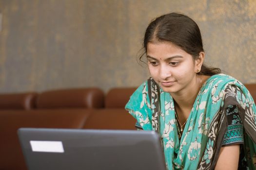 Smiling indian girl student or employee busy on laptop sit at home in casual dress, happy woman studying, e-learning, using online software or technology app for work, education concept.